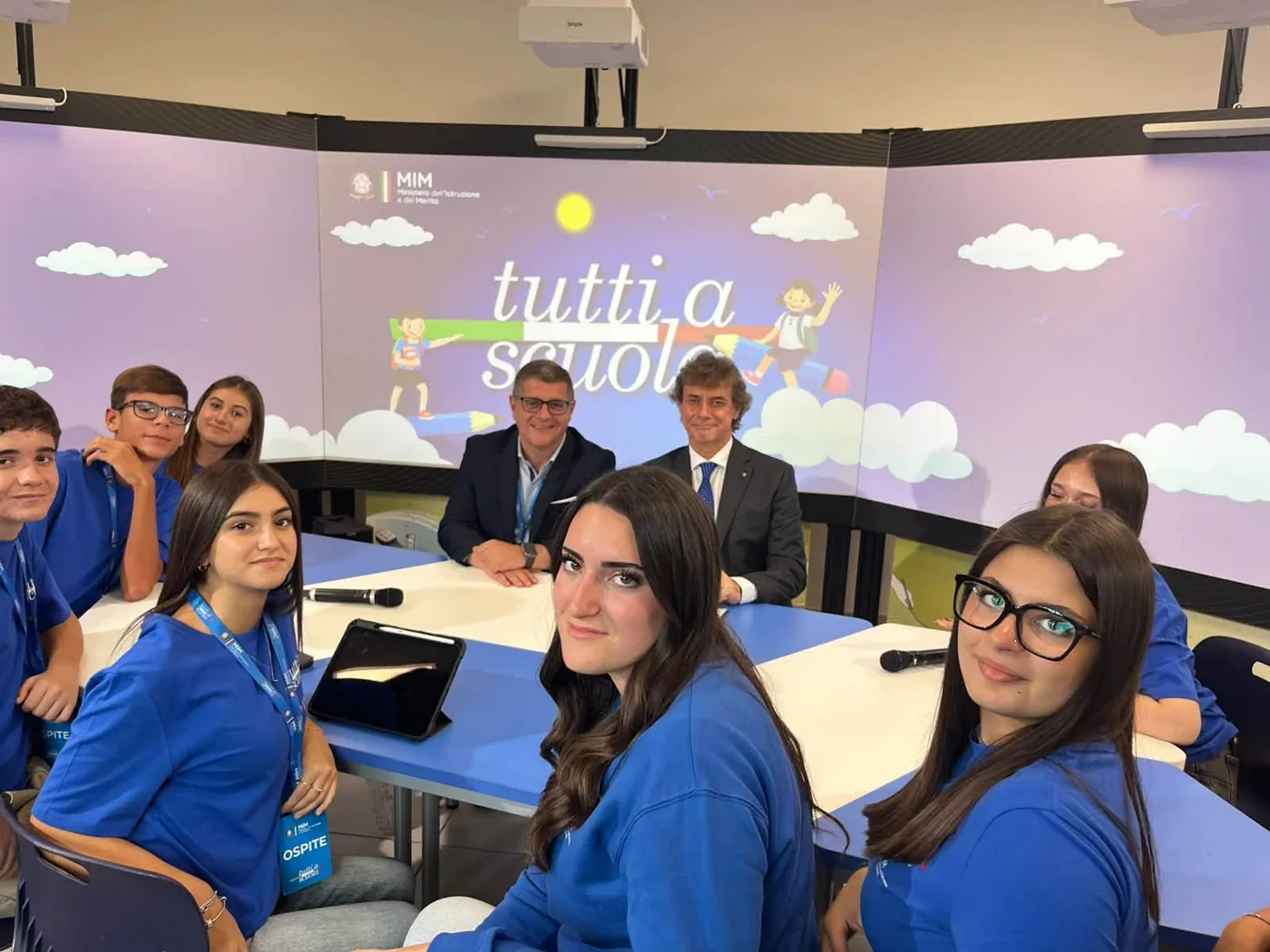 Gruppo di studenti con magliette blu seduti attorno a un tavolo nell’aula immersiva; sullo sfondo la scritta «Tutti a scuola» e il logo del Ministero dell’Istruzione e del Merito.
